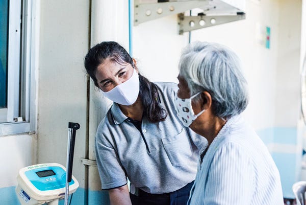 In a healthcare setting, a health worker wearing a mask is leaning forward and smiling at a seated patient that is also wearing a face mask.