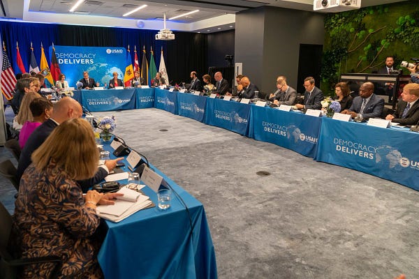 Secretary of State Antony Blinken sits at a rectangular table that are connected to form a "U" shape with blue table cloth that says "Democracy Delivers". Leaders sit around the table to discuss how democracy can deliver.