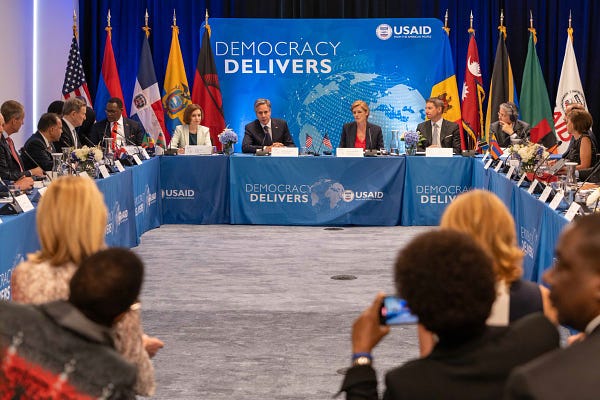 Secretary of State Antony Blinken sits at a rectangular table that are connected to form a "U" shape with blue table cloth that says "Democracy Delivers". Administrator of the United States Agency for International Development Samantha Power sits on the left of Secretary Blinken wearing a red shirt with a black blazer. There is a blue poster behind the tables that also says "Democracy Delivers" .
