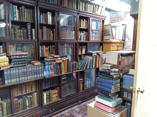 A dark wood bookshelf full of old books, with more in precarious stacks aroud it, in a low-ceilinged nondescript room with bits of brick wall and faded antique rug visible