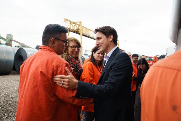 Prime Minister Justin Trudeau is standing outdoors and shaking hands with a worker. Other people are standing behind them.