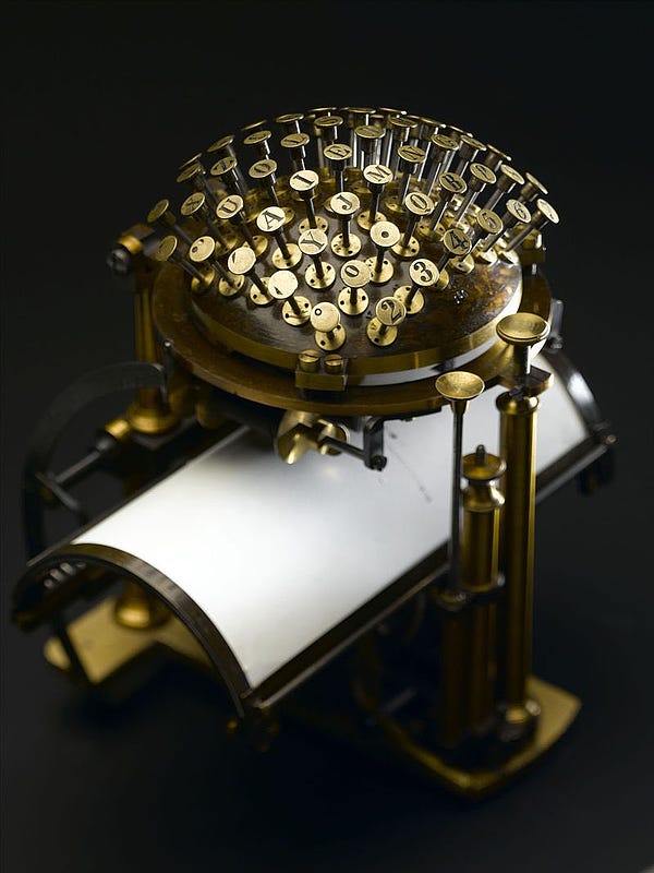 A mechanism holding a paper on a curved cylindrical mount surmounted by a ludicrous brass hemisphere covered in brass punches with letters and numbers on it like a pincushion but for writing