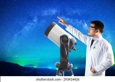 A scientist in a lab coat points at a space thing. He is not using the telescope in front of him.