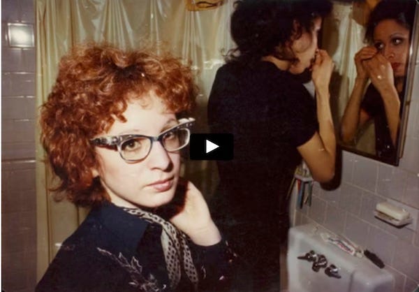 Color still from "All the Beauty and the Bloodshed" showing a young Nan Goldin. She is in a bathroom. Another woman is in the background applying makeup in the mirror.