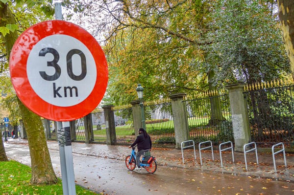 A close-up of a red-and-white 30 km/h speed limit sign in Brussels. A cyclist passes by on the street in the background.