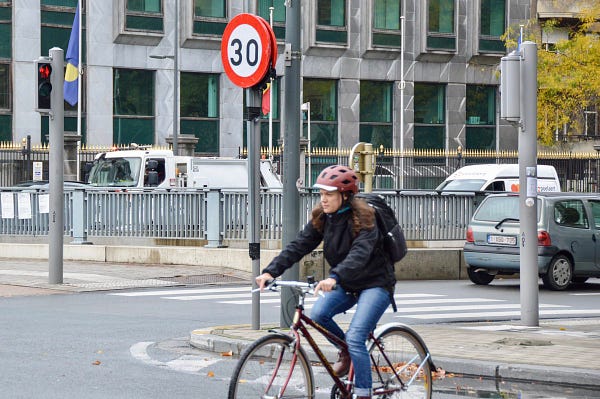 A red-and-white 30 km/h speed limit sign in Brussels. A cyclist passes by on the street in the foreground.