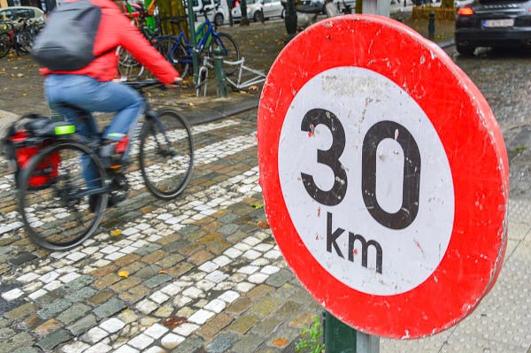 A close-up of a red-and-white 30 km/h speed limit sign in Brussels. A cyclist passes by on the street in the background.