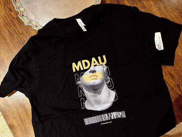 A black T-shirt has the word “MDAU" printed over what appears to be a photo of a Roman or Greek sculpture. The T-shirt is draped over a wooden table. A silver platter is visible on the table.