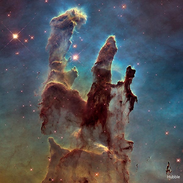 Alt text from NASA: Hubble’s visible-light view of a star-forming region, the Pillars of Creation, shows darker pillars that rise from the bottom to the top of the screen, ending in three points. The background is opaque, set off in yellow and green toward the bottom and blue and purple at the top. A handful of stars of various sizes appear.