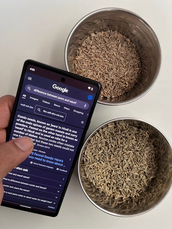 A phone with the Google app open, with ‘difference between jeera and saunf’ written in the Search box, with two bowls filled with jeera and saunf in the background.