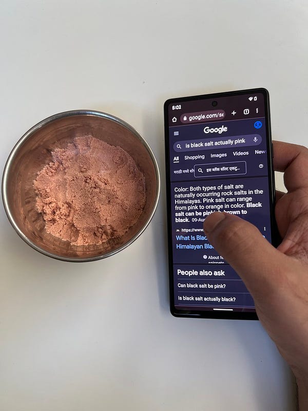 A phone with the Google Chrome app open, with ‘is black salt actually pink’ written in the Search box, with a bowl full of black salt in the background.