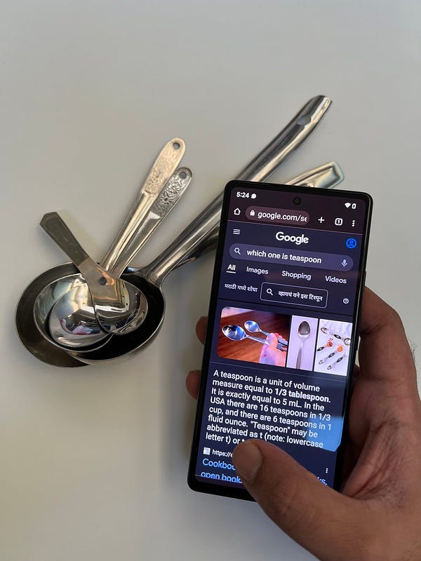 A phone with the Google Chrome app open, with ‘which one is teaspoon’ written in the Search box, with a bunch of spoons of various sizes in the background.