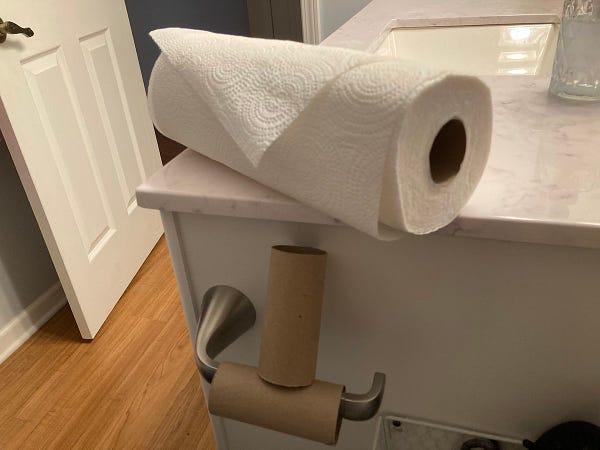 one empty toilet paper roll on a holder in the bathroom. another empty roll is propped on top of it. directly above that on the sink counter is a roll of paper towels that appears to have been recently used. wrongly.