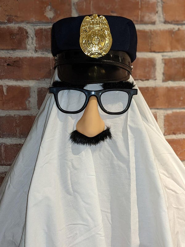 A white sheet covers a figure who is wearing joke glasses (with furry eyebrows and mustache and nose attached), with a police hat on. The figure stands in front of a red brick wall facing the camera. 