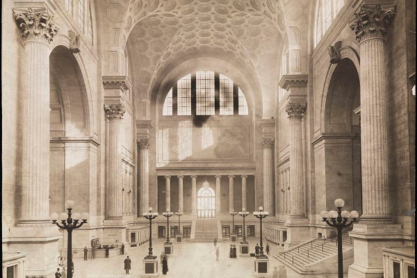 Black and white photo of the main waiting room in Penn Station around when it opened in 1910. It’s mostly made of stone with huge vaulted ceilings held up by corinthian columns. 