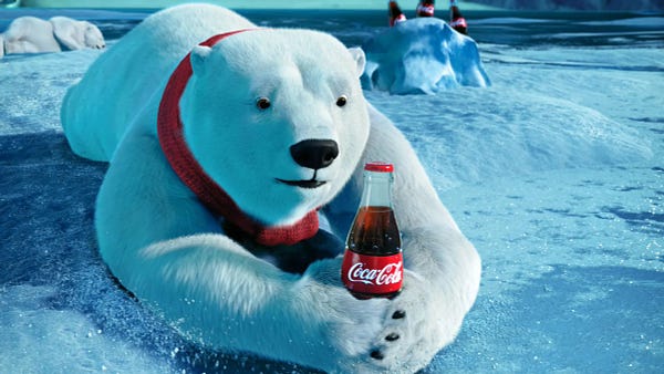 Image of one of the Coca-Cola polar bears wearing a red scar, lying in the snow, and looking at a Coke bottle