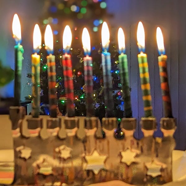 A fully rainbow candle lit menorah in front of a rainbow lit Christmas tree 