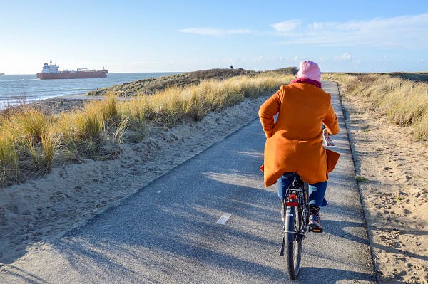 A woman in an orange wool coat and pink winter hat rides a black electric bike on a separated cycle path in rural Zeeland, the Netherlands.