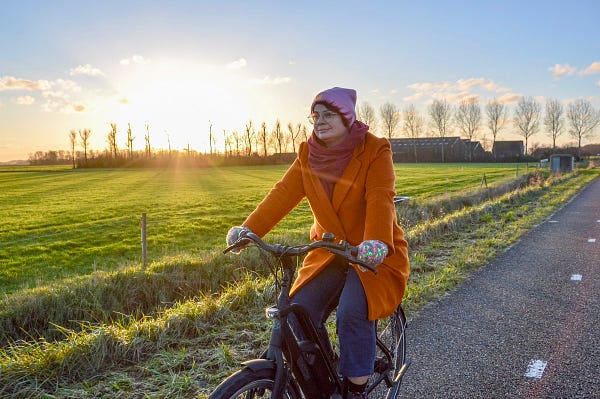 A woman in an orange wool coat and pink winter hat rides a black electric bike on a separated cycle path in rural Zeeland, the Netherlands.