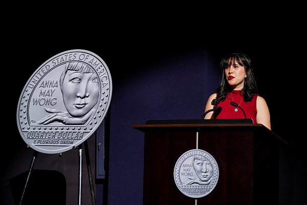 Writer Katie Gee Salisbury speaks about Anna May Wong's legacy (in red dress standing behind lecturn -- coin blow up of the Anna May Wong quarter on the left, smaller image of the quarter placed on the lecturn).