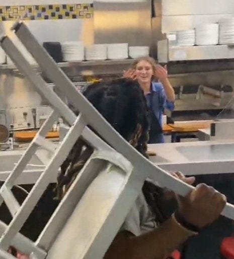 In the foreground a woman raises a metal chair to throw it at a Waffle House employee in the background. The Waffle House employee holds her hands up as if to say “bring it on”.