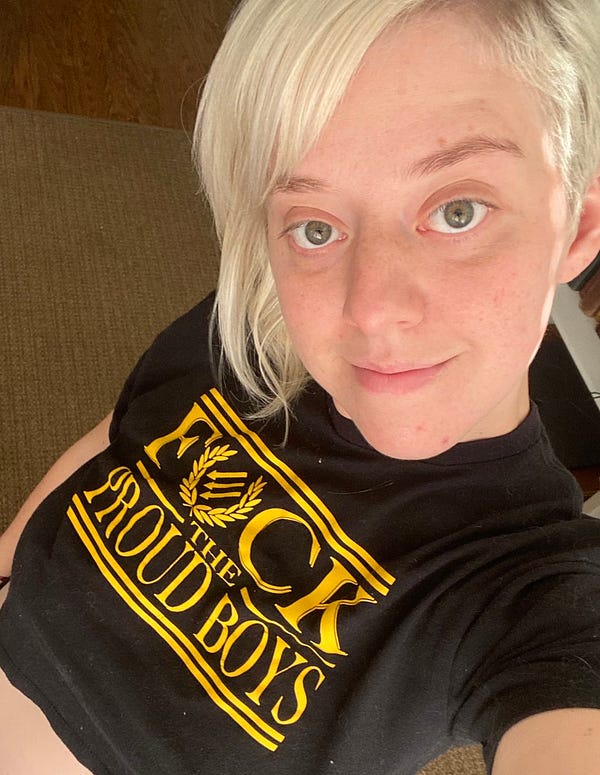 An overhead selfie of Melissa. She has a bright blonde side shave that is short on one side, and bright blue eyes. She is wearing a black and yellow shirt that says FUCK THE PROUD BOYS