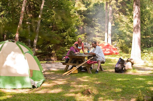 Three adults sit at a picnic table surrounded by camping equipment in front of a tent at a campground on a sunny day.