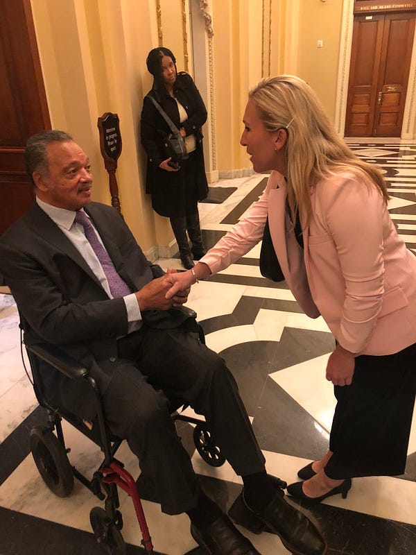 Left: Jesse Jackson, a black man in a suit seated in a wheelchair. 
Right: Marjorie Taylor Greene, a blonde white woman in a white suit jacket, a sequined black shirt and black pants, shaking his hand.