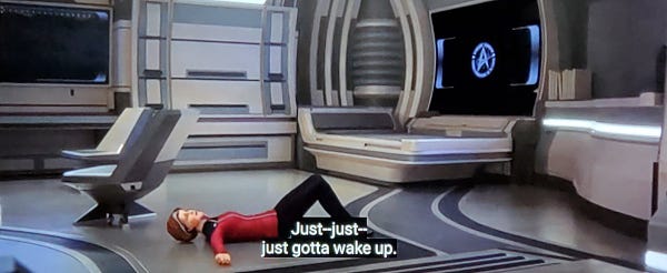 Scene from Star Trek Prodigy, personal quarters shown with a starship/sterile version of a bed and two chairs shown in background and a video screen on the wall with a delta shield screensaver image. Admiral Janeway is laying on the floor on her back, arms outstretched. Closed caption reads, "Just--just--just gotta wake up."