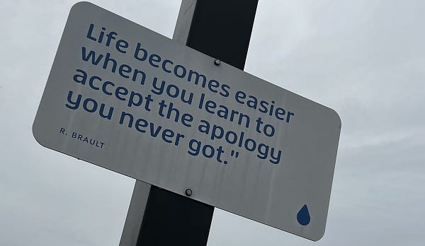 A white metal sign with blue lettering and a quote that reads, "Life becomes easier when you learn to accept the apology you never got," citation is to R. Brault