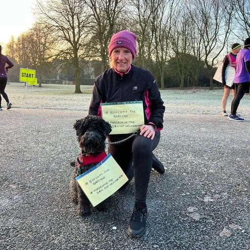 Sue Roberts, with her dog poppy, at Birkenhead parkrun. She is wearing a t-shirt with a slogan pinned to it which says 'Bin the barcode'