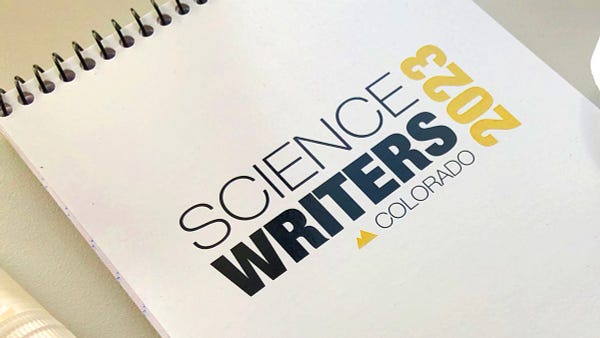 Horizontal photo of a close up of a reporters spiral notebook placed diagonally on a table, with the Science Writers 2023 Colorado logo, which includes a small mountain peak motif.