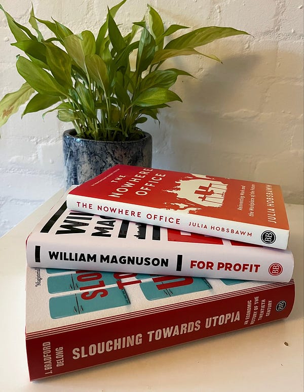 A stack of 3 hardcover books including Slouching Towards Utopia, For Profit and The Nowhere Office, sitting on a white desk against a white brick wall. There is a houseplant in the background.