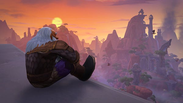 A close-up view of the dragon Veritistrasz in his dwarf visage form. The gray-haired dwarf wears simple clothing and sits on a stone ledge overlooking the mountains, canyons, and ruins of the Waking Shores during an orange sunset. 