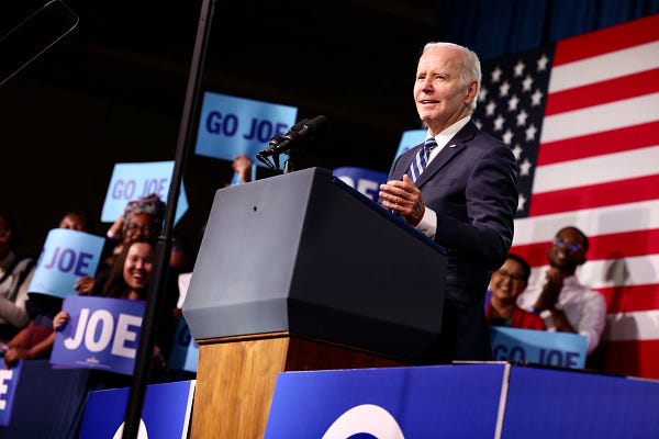 President Joe Biden speaking to an audience. Next to him are people holding signs that read "JOE" and "GO JOE"