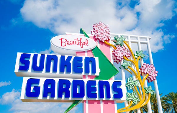 Looking up at a colorful, vintage-style sign that reads “Beautiful Sunken Gardens” on a bright, sunny day.