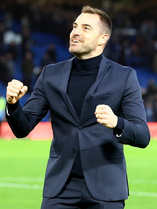 A smiling Rubén Sellés, wearing a dark-coloured turtleneck under a navy suit, clenches both fists and looks upwards as he celebrates in front of Southampton’s fans after the team’s 1-0 win at Chelsea.
