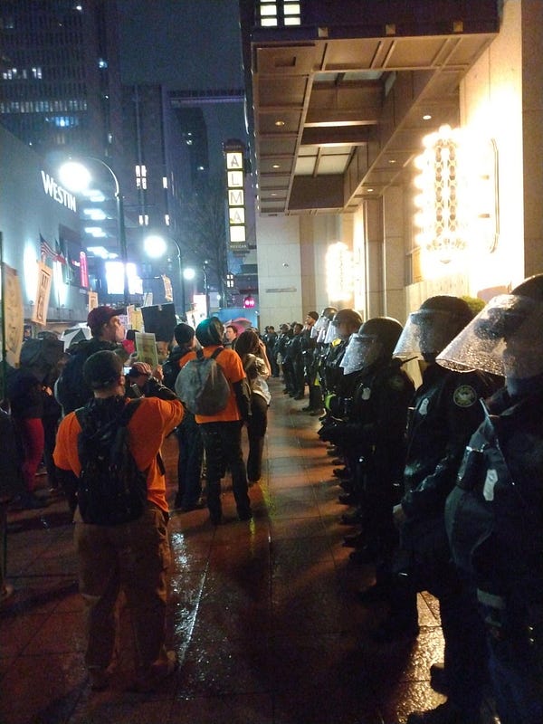 A police riot line stands in front of a building. Individuals with orange shirts working as cop watch stand in front of them, behind cop watch stands a large protest.