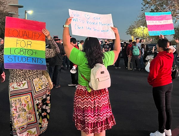 Three people holding signs, a crowd can be seen behind them with several wearing cat ears.

One sign is rainbow colored, reading “equality for LGBTQ+ students”

Another reads “We [HEART] our WESD Board members (Just like Jesus would)

Another, in the colors of the trans flag, reads “ 1.8 million queer youth seriously considered suicide in the past year. STOP KILLING KIDS.”