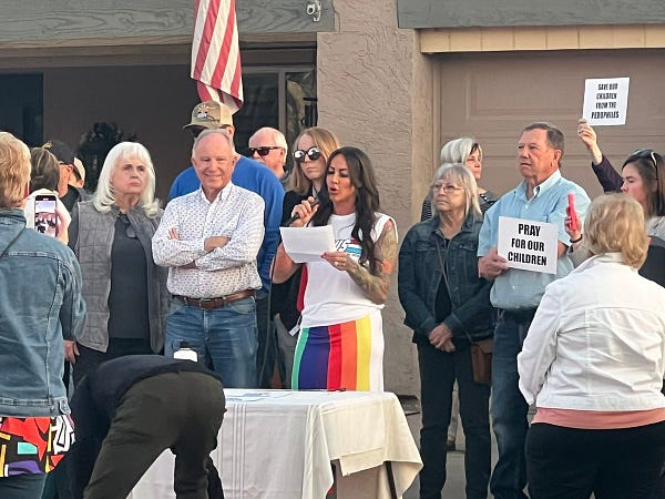 Graham in a bright colorful rainbow skirt, reading off a piece of paper into the mic.

Around her around others in Kern’s group, including two people holding signs reading “pray for our children” and “save our children from the pedophiles.”