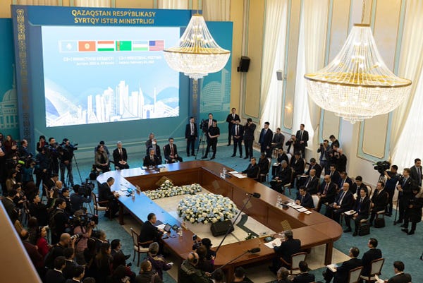 Secretary Blinken and C5 officials sit around a hollow rectangular table in a large room. A large screen shows the flags of the participating countries and members of the press take photos.  