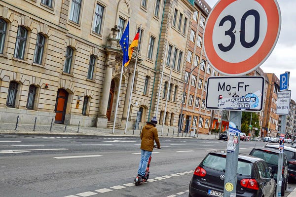 A man on a kick scooter in winter attire travels past a large “30 km/hr Zone” sign in Berlin.