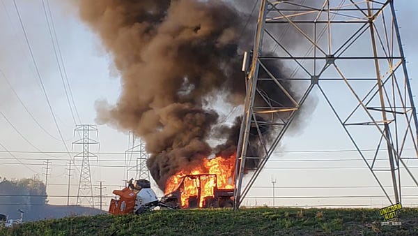 A police vehicle on a green hill under power line towers burns brightly in large hot orange and red flames with a large plume of smoke rising.