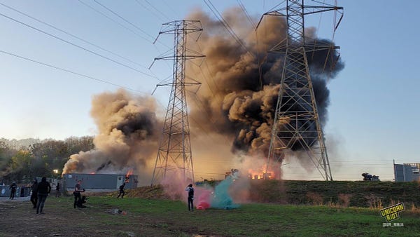 Colored flares erupt as vehicles and other parts of the Cop City construction outpost burn up as a masked crowd overtakes the area.