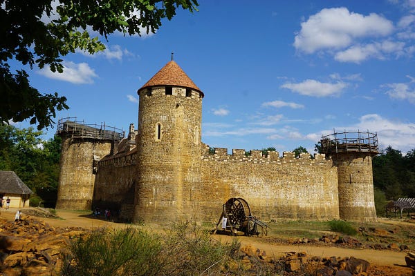 A photo of a medieval style castle under construction in a sunny French forest, part of a giant hamster wheel used to operate a manual crane sits in the foreground along with scaffolding on the incomplete towers.