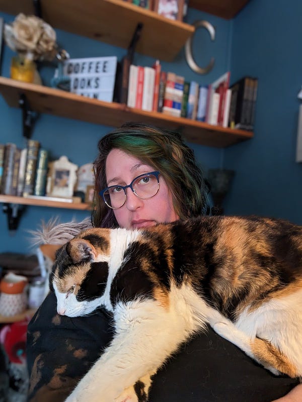 A calico cat lays on a white woman's chest. The woman is wearing glasses and has multi-colored hair. She is sitting in a desk chair and there are bookshelves behind her.