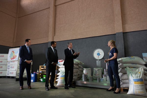 Secretary Blinken and Ethiopian Minister of Finance Ahmed Shide speak with a woman who is weighing sacks of grain inside a warehouse. 