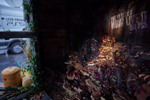 A wider shot of the Cordyceps infected person on a sofa, their skull and body is covered in fungi and spores. To the left of the photo you can see a white van with a satellite dish on top of it with the words "Live from PS5" printed on the side.  They were located in BOXPARK Shoreditch in London. 