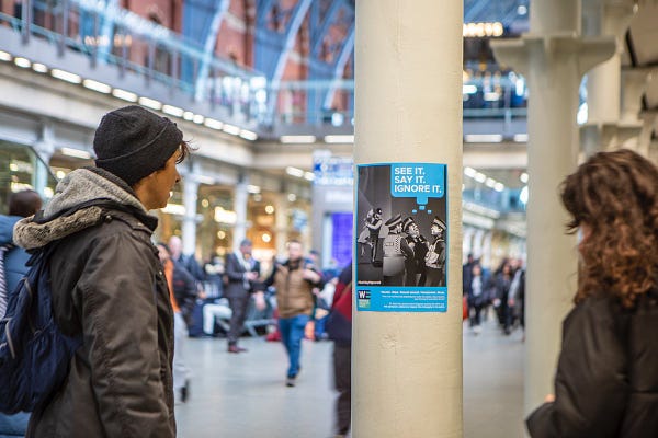 An image of two members of the public looking at a poster of a black and white illustration of police officers ignoring a woman being attacked by a fellow police officer. The words, "See it, say it, ignore it" are on the top right of the poster, and the poster is in a busy train station. 