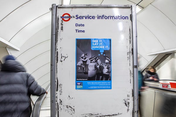 A poster of a black and white illustration of police officers ignoring a woman being attacked by a fellow police officer. The words, "See it, say it, ignore it" are on the top right of the poster, and the poster is attached to a service information board in a tube station.